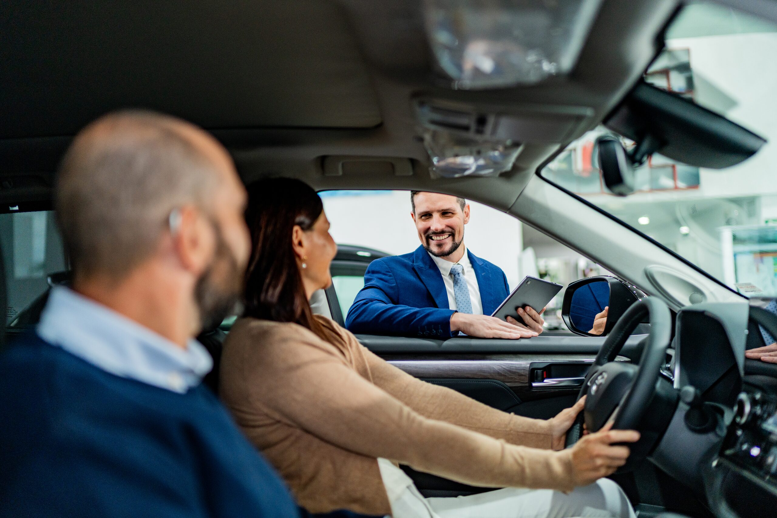 A salesperson with a tablet leans into the driver's window as a couple sits in a new car