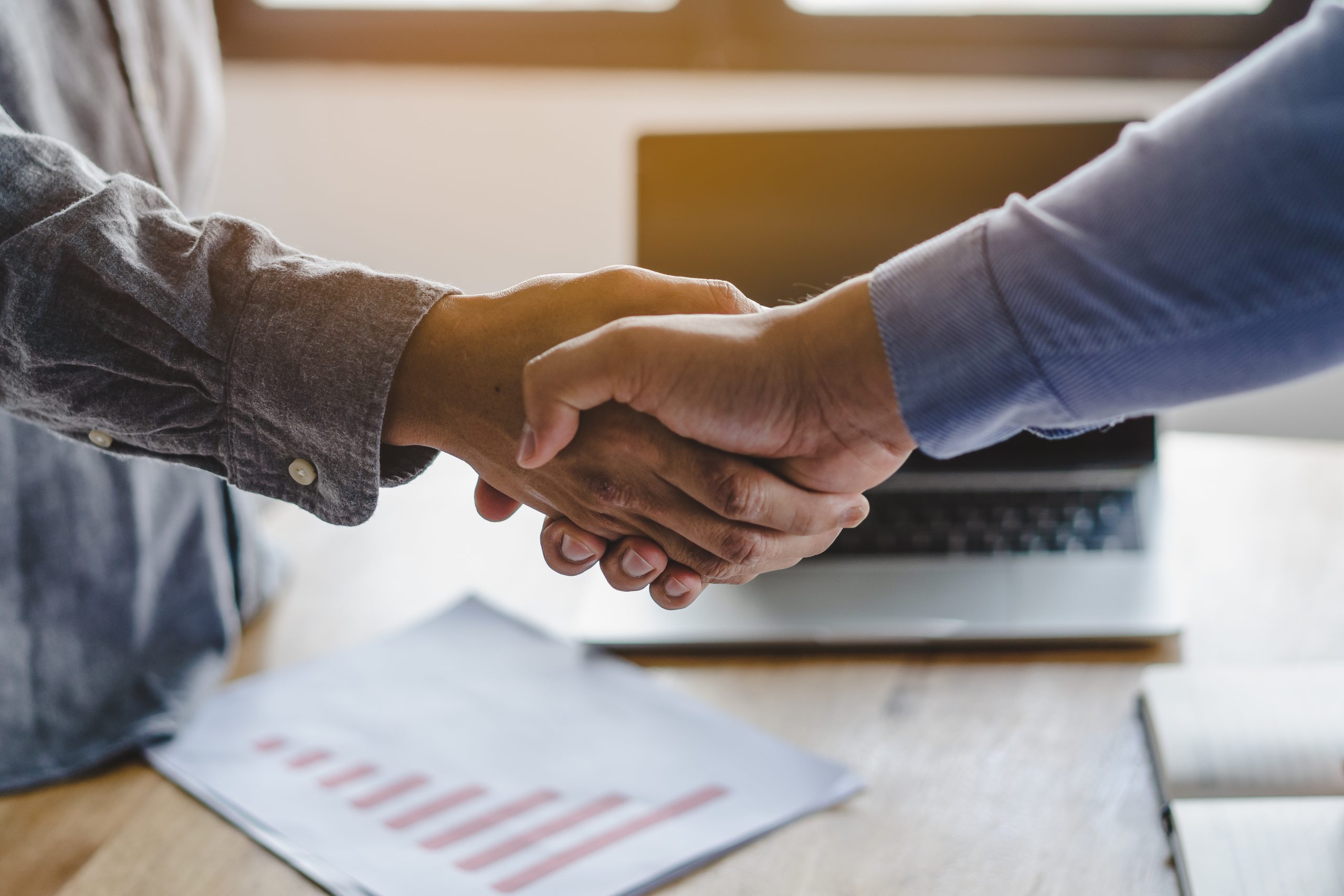 An advisor and a client shake hands over a desk with a laptop and a printed chart
