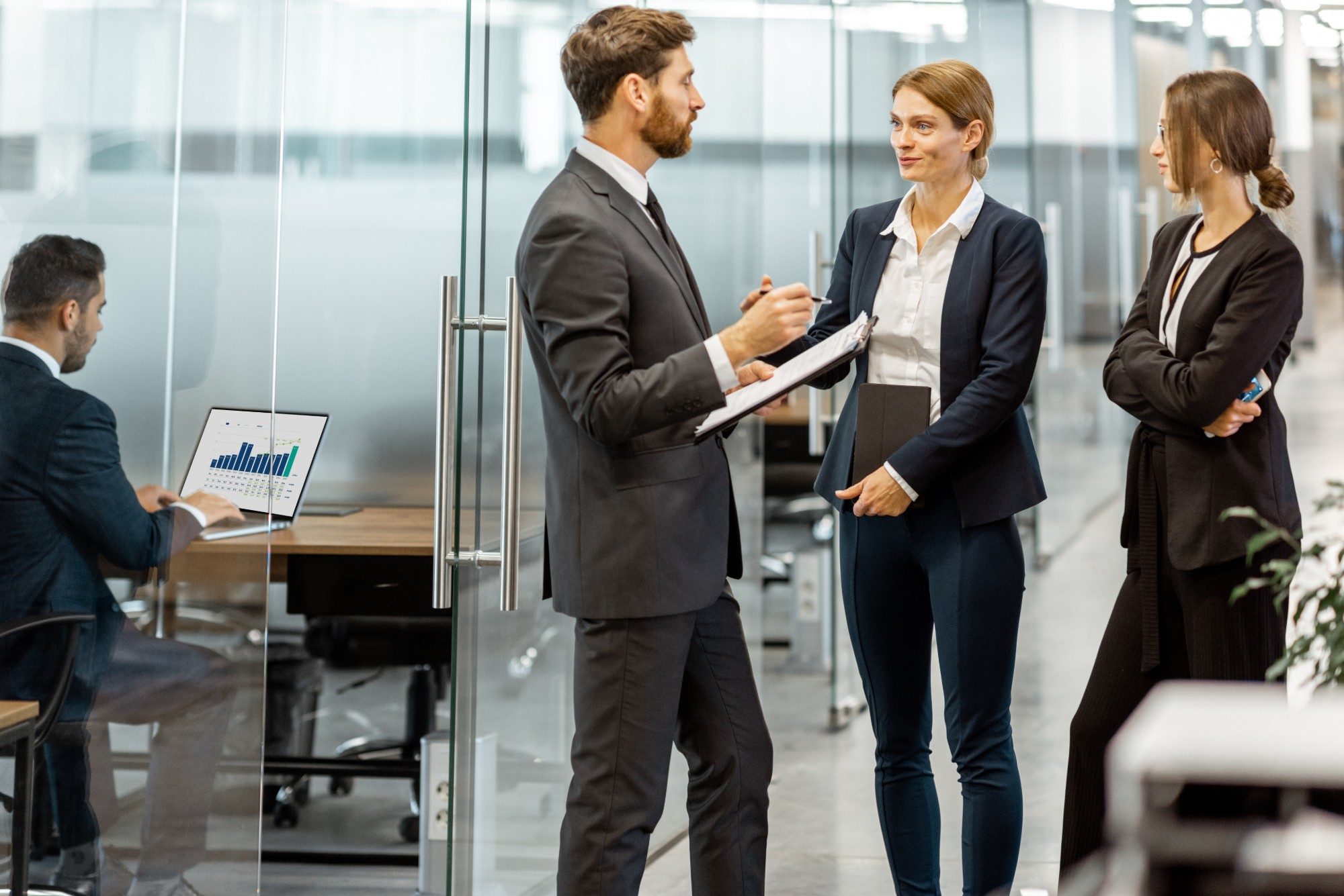 An advisor reviews notes on a clipboard with two colleagues in a glass-walled office
