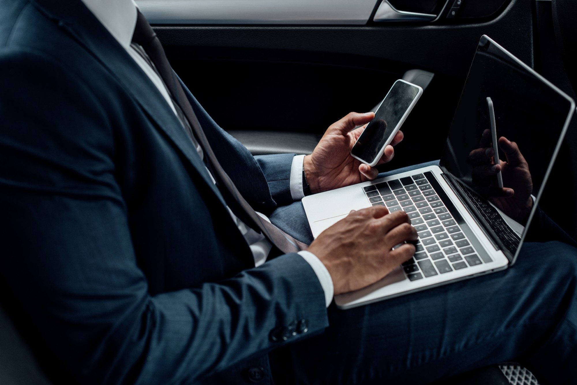 A rep updates his CRM on a laptop and phone in the back of a car between visits