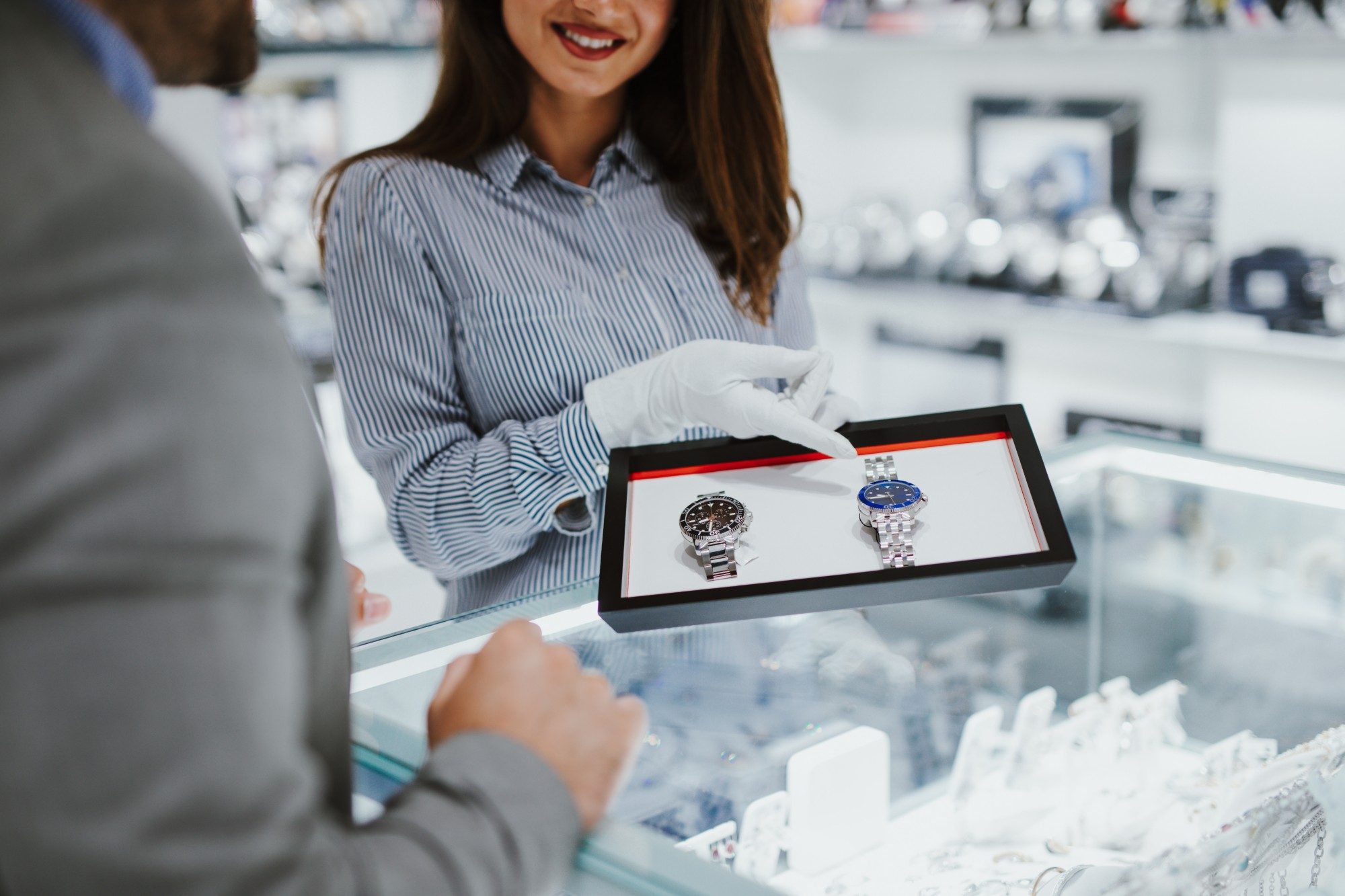 A seller in white gloves shows a customer two watches on a tray at the counter