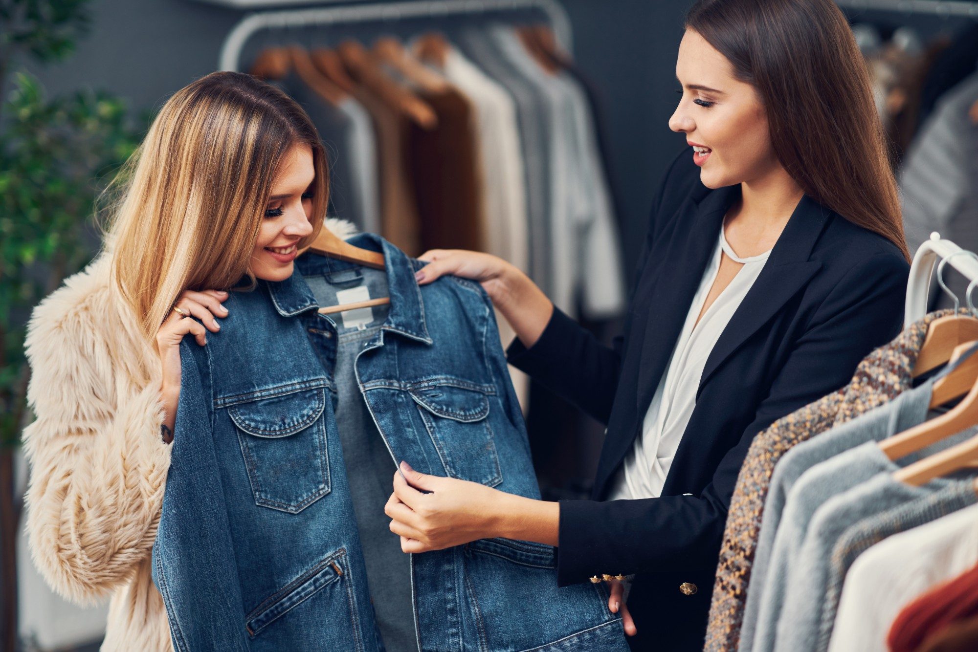 A seller holds up a denim jacket for a smiling customer in a clothing boutique